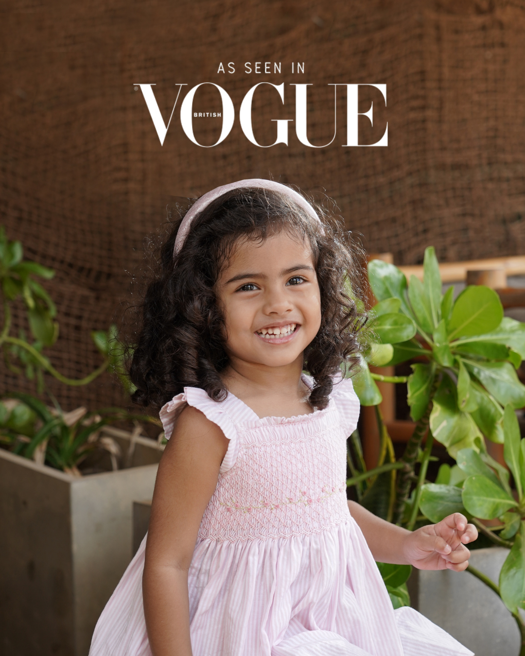 Toddler girl in a pink dress sitting outdoors with plants as seen in British Vogue.